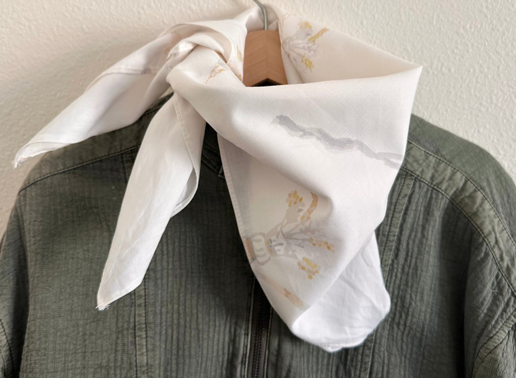 A white silk scarf with a delicate gray and yellow floral print, tied around a wooden hanger and draped over a textured olive green shirt against a neutral wall.