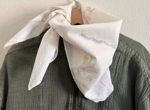 A white silk scarf with a delicate gray and yellow floral print, tied around a wooden hanger and draped over a textured olive green shirt against a neutral wall.