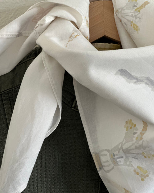 A close-up of a white scarf with a delicate gray and gold botanical pattern, draped over a dark charcoal gray ribbed knit top.
