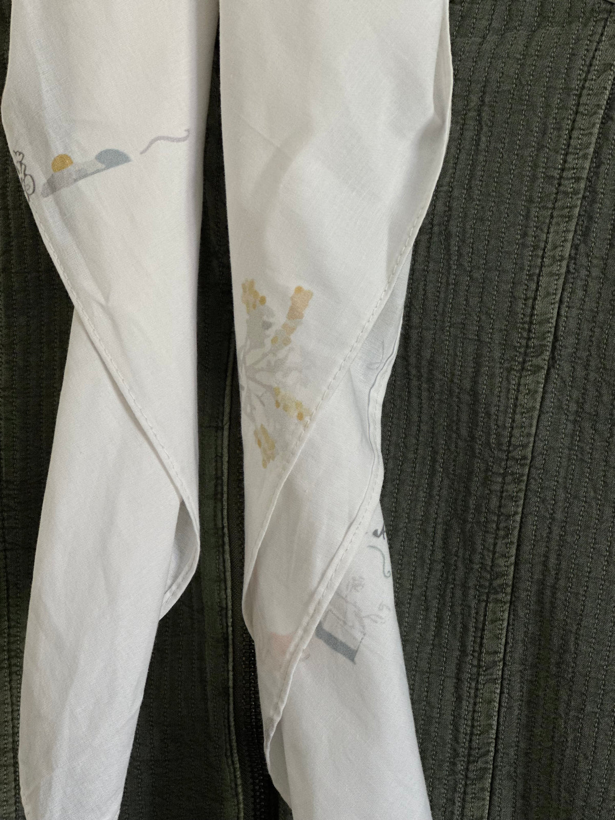 A close-up of a white fabric garment, likely a skirt or dress, featuring a subtle gold floral pattern and light blue and gray abstract accents, draped against a dark textured olive green fabric background.