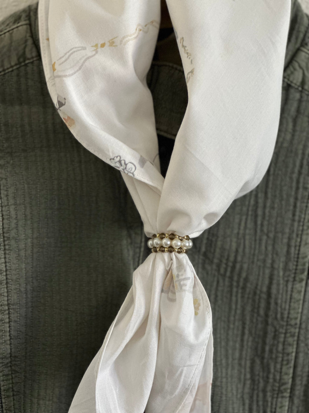 A close-up of a white silk scarf with delicate gold and gray patterns, tied around the neck of a textured dark gray shirt. The scarf is secured at the collarbone with a decorative pearl and gold chain tie.