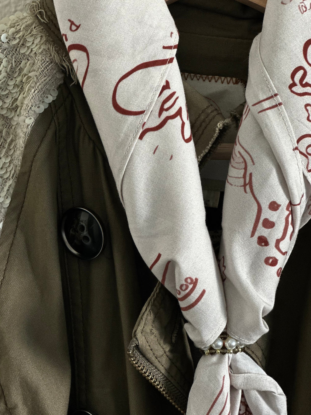 A close-up of a cream scarf with a rust-red abstract line pattern tied around a dark olive green jacket on a wooden hanger, next to a textured beige sequined garment.
