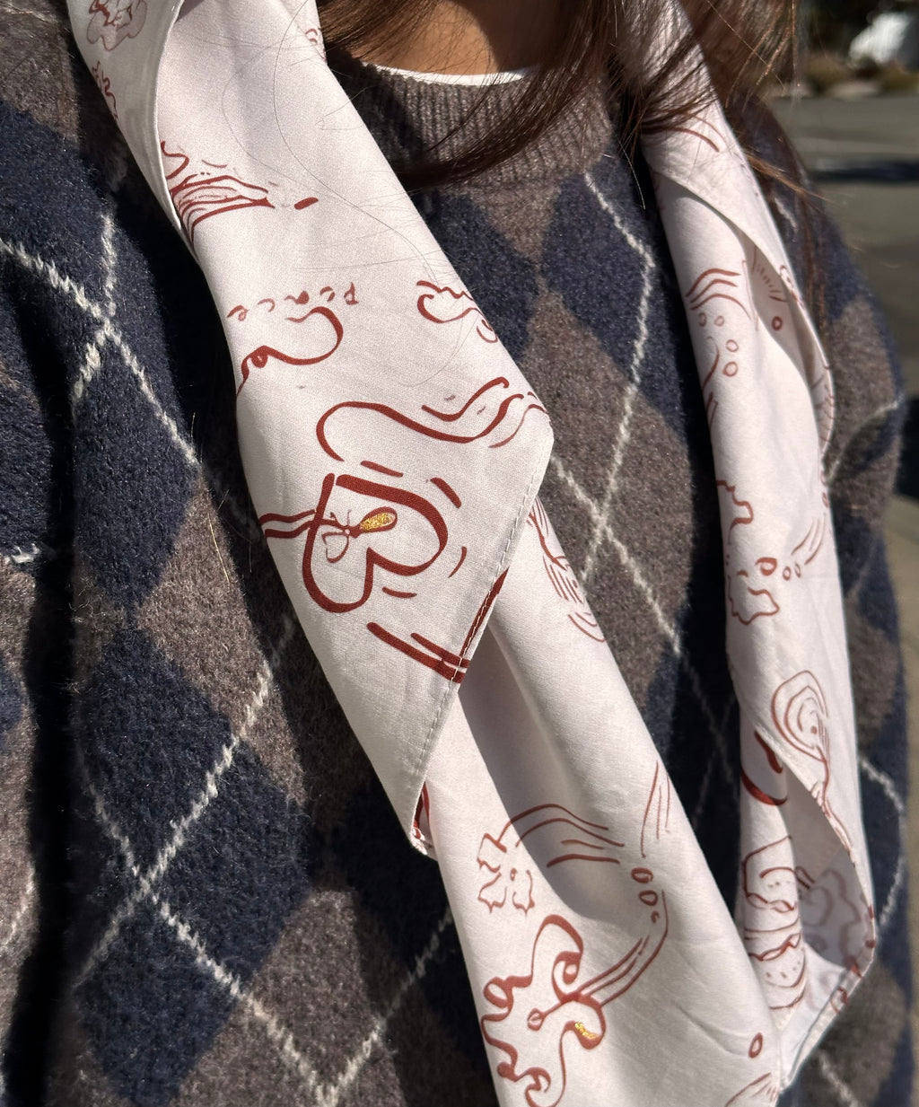 A close-up of a person wearing a white scarf with red abstract line drawings and the word 'Peace' printed on it, layered over a navy and gray argyle sweater.