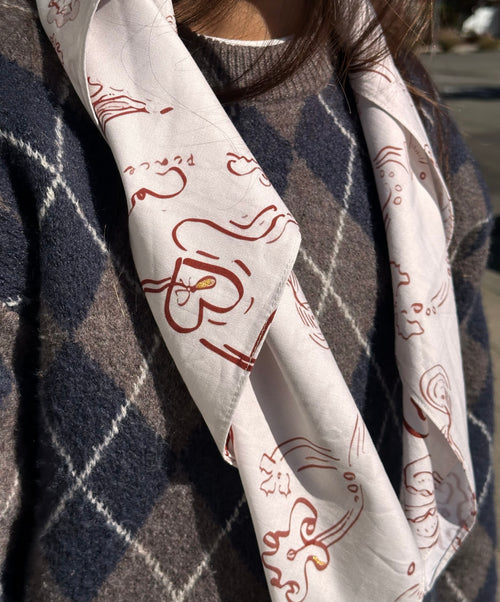 A close-up of a person wearing a white scarf with red abstract line drawings and the word 'Peace' printed on it, layered over a navy and gray argyle sweater.