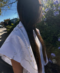 A woman outdoors wearing a white scarf with gold celestial line art and small gold stars, photographed from the side in bright sunlight.