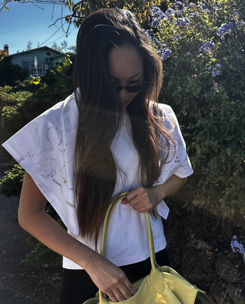 A woman outdoors wearing a white short-sleeve top with gold floral embroidery, holding a light yellow handbag. She is wearing dark sunglasses, with green foliage and purple flowers in the background.